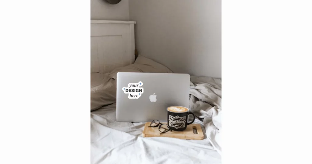 a sticker mockup on a Macbook, with a coffee cup and glasses on the bed